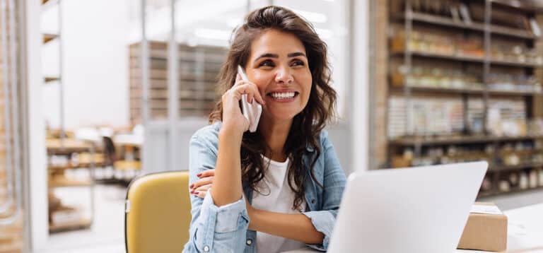 woman with brown hair and brown eyes smiling while on the phone sitting in front of her laptop