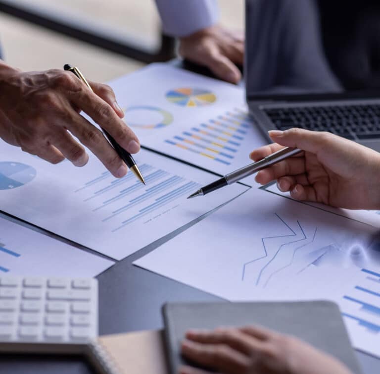 close up of hands with pens and printed out charts and graphics next to open laptop