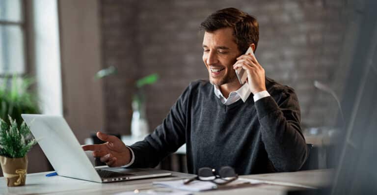 man at desk smiling while on phone and looking at open laptop