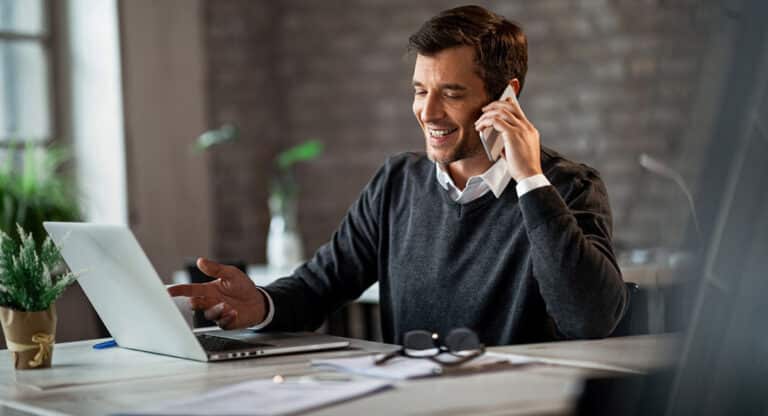 man at desk smiling while on phone and looking at open laptop