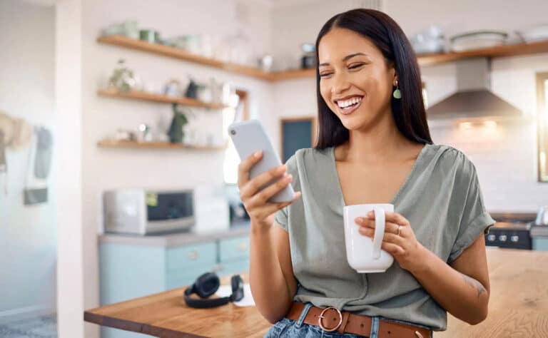 woman smiling into phone holding white coffee cup standing in modern kitchen
