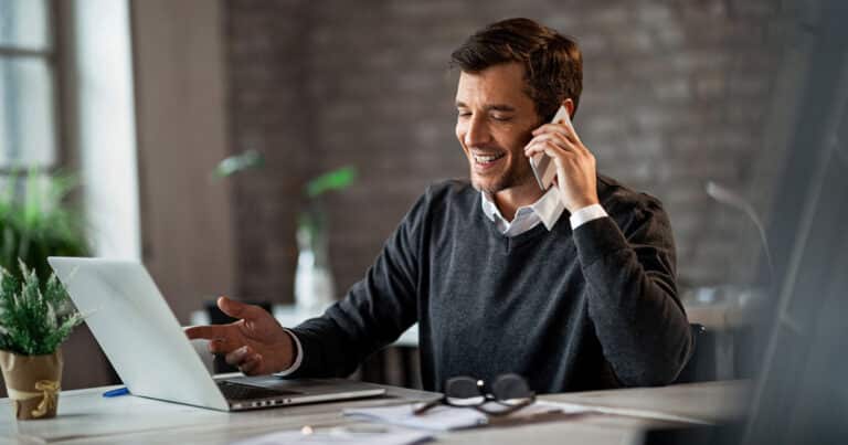 man at desk smiling while on phone and looking at open laptop