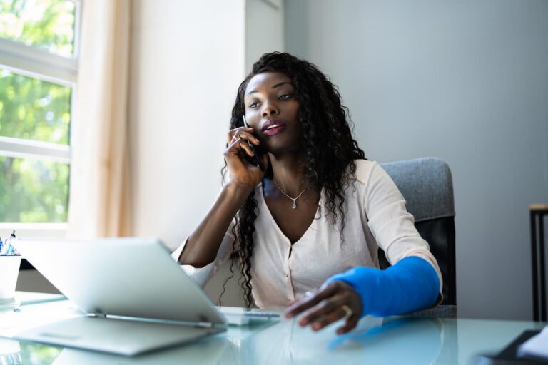 woman on phone talking and in front of laptop at desk with blue cast on arm