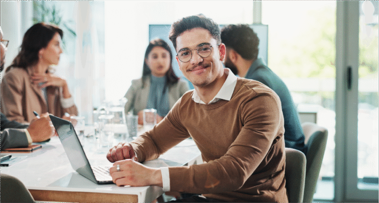 Man with glasses and sweater smiling in front of laptop while in a boardroom setting with four colleagues.
