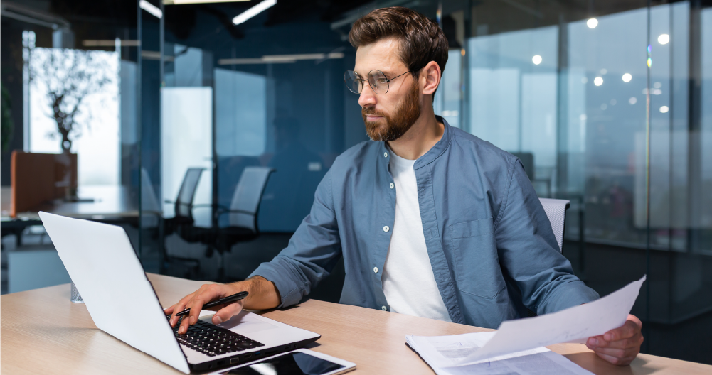 A man financier, auditor sits in the office . He holds documents, checks accounts. types on a laptop.