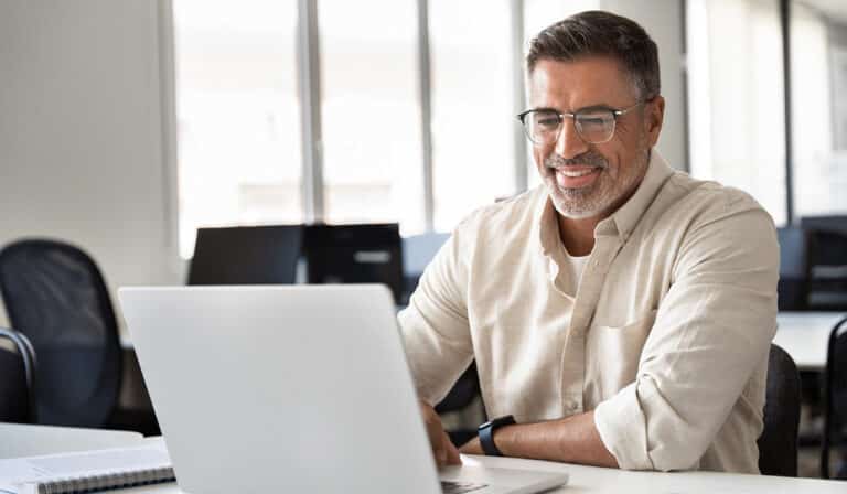 man with glasses and beard smiling in front of laptop notebook and phone