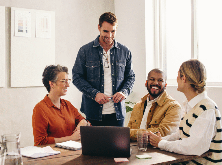 Four adults collaborating at table looking at laptop while smiling.