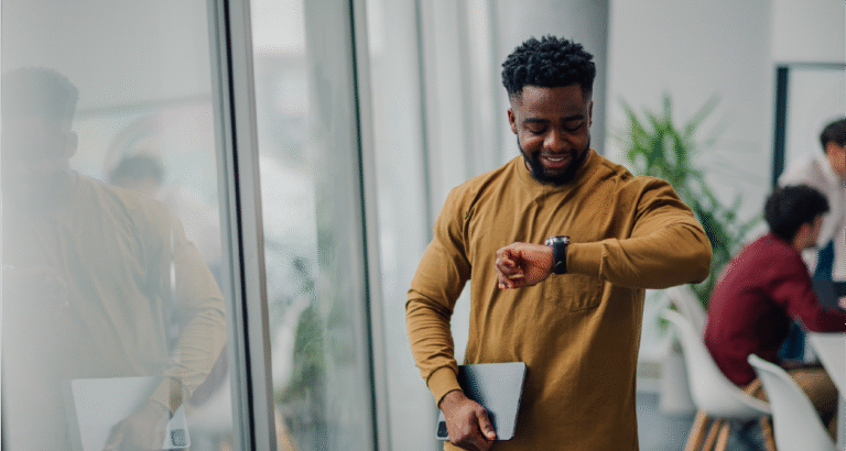 A man standing in the office in front of windows holding a tablet and is looking at his watch while smiling.