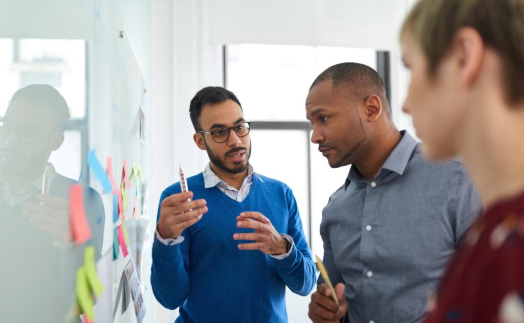 coworkers looking at a whiteboard with sticky notes as one team member discusses something on the board