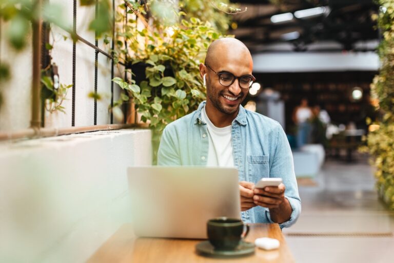 Man sits in a cafe, casually dressed and smiling while using his smartphon