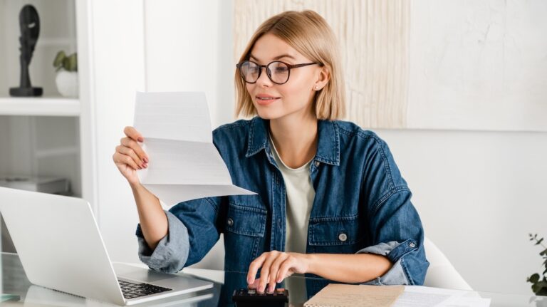 young woman looking at a bill in front of her laptop while her fingers are on a calculator.