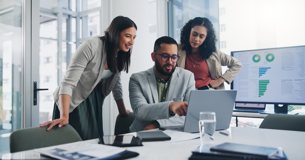 team working together in front of a laptop with data showing on a tv screen in the background