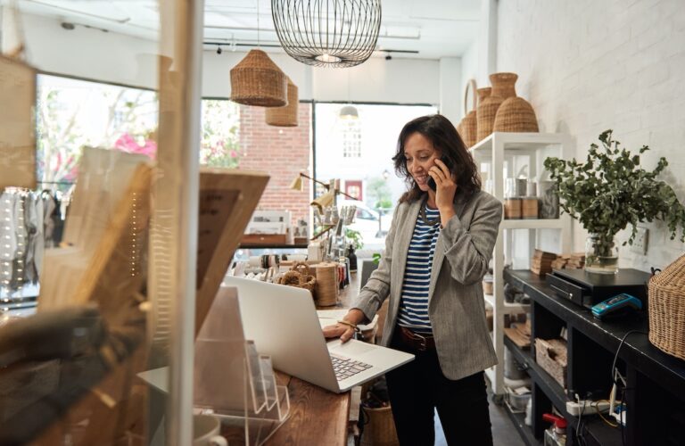 young woman standing behind a counter in her stylish boutique working on a laptop and talking on a cellphone