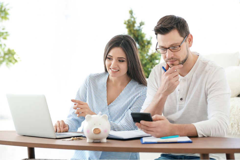 a couple sitting at a desk in front of a laptop while the man is on his phone. a piggy bank sits in front of them while discussing finances