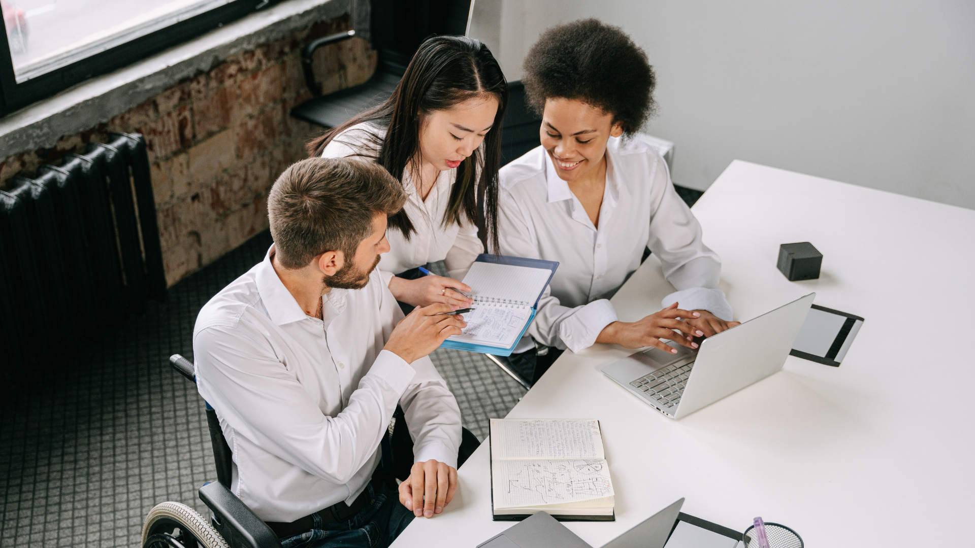 Three coworkers, two female and one male, sit around each other reviewing a document one lady is holding