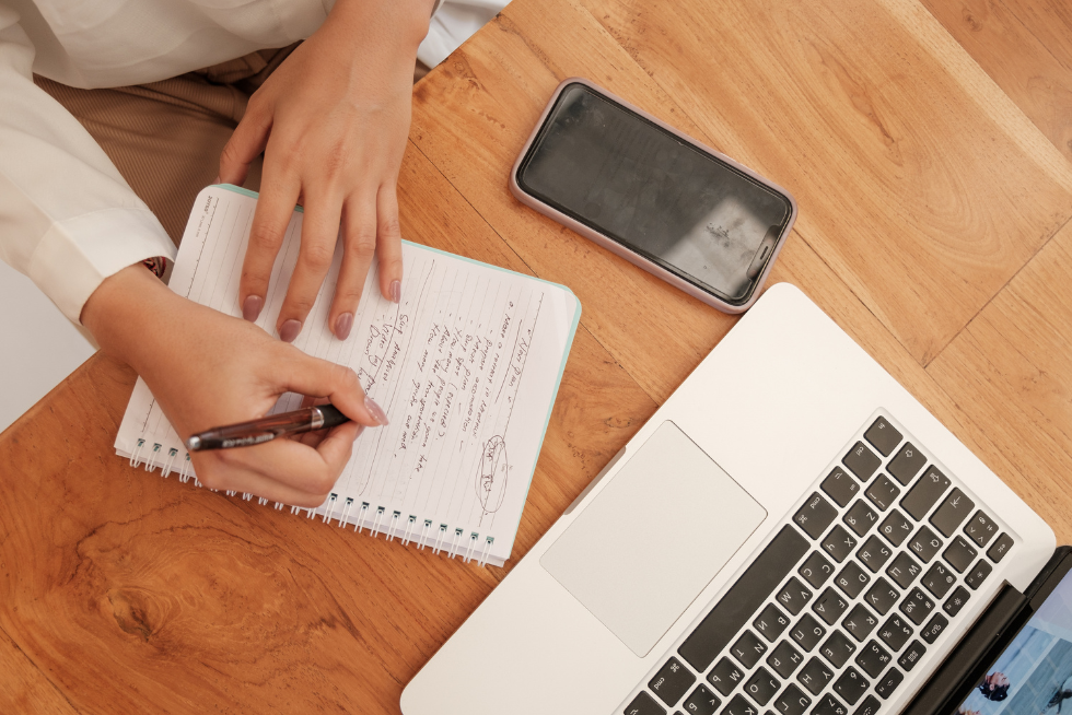 Photo from above of a laptop, cellphone, and a woman's hands writing notes in a small spiral notebook