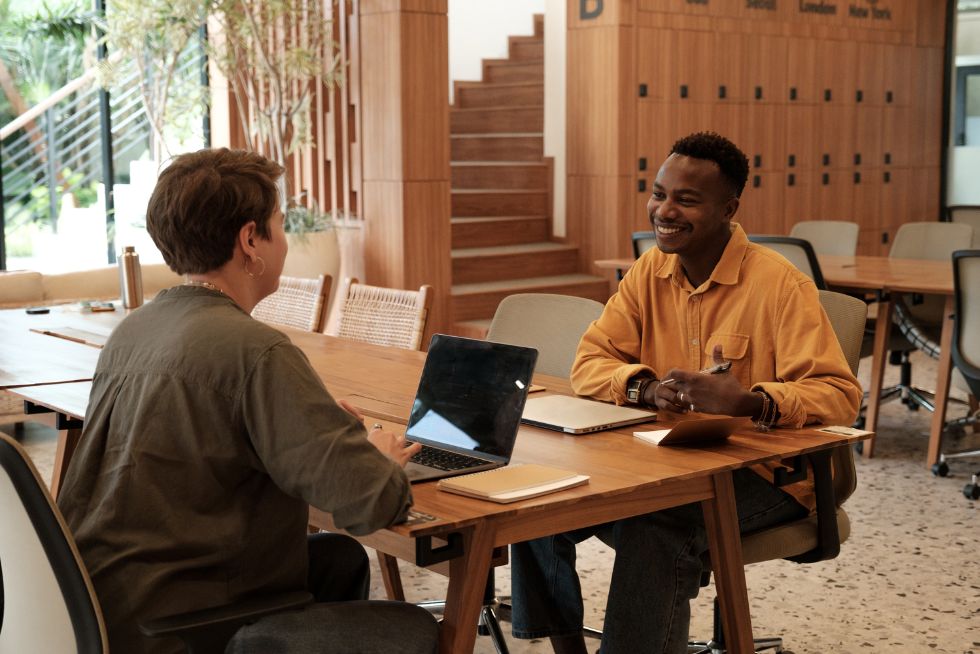 a man and a women sitting at a table talking with their laptops in front of them