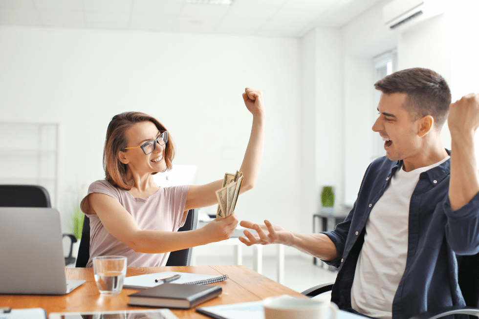 Two coworker, one female wearing glasses and one male, celebrating while the female holds money in her hand