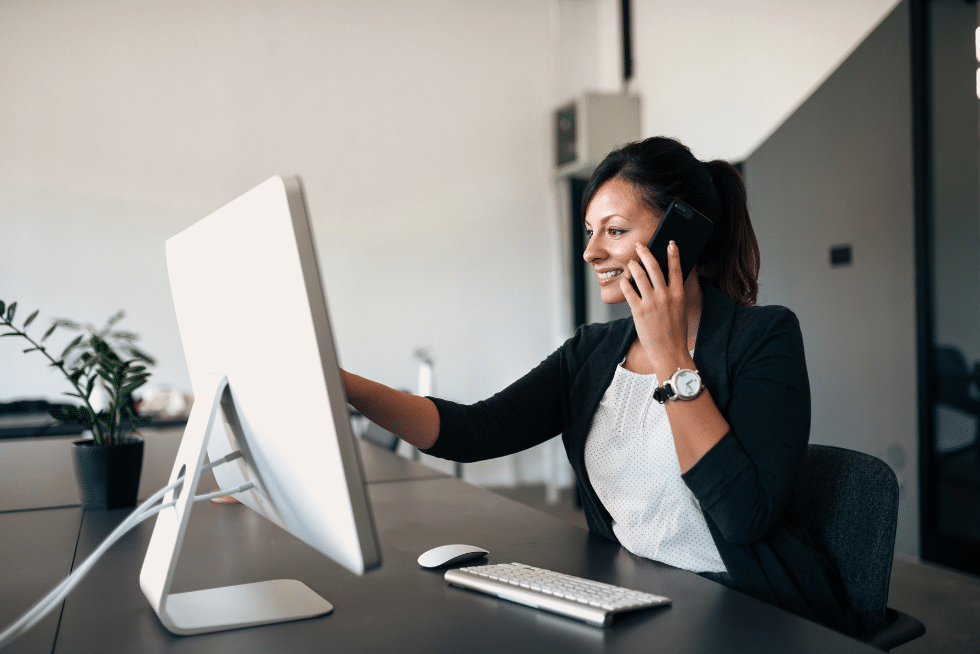 woman sitting at a desk, speaking to someone on her cellphone while sitting in front of a desktop monitor.