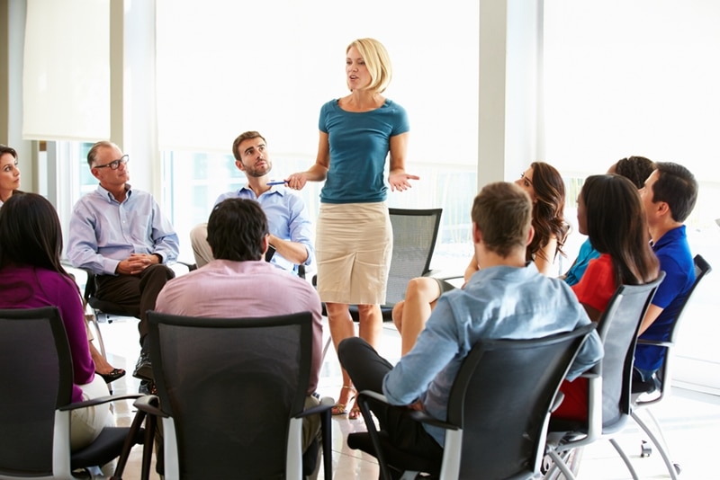 A group of employees having a meeting sitting down in a circle and a women standing up and speaking
